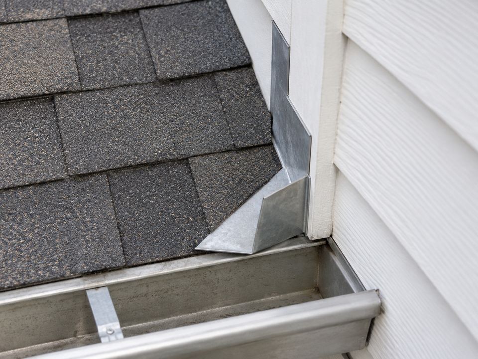 A bent metal diverter at the lower end of a roof-to-wall junction, with its angled wing projecting away from the wall over a gutter.