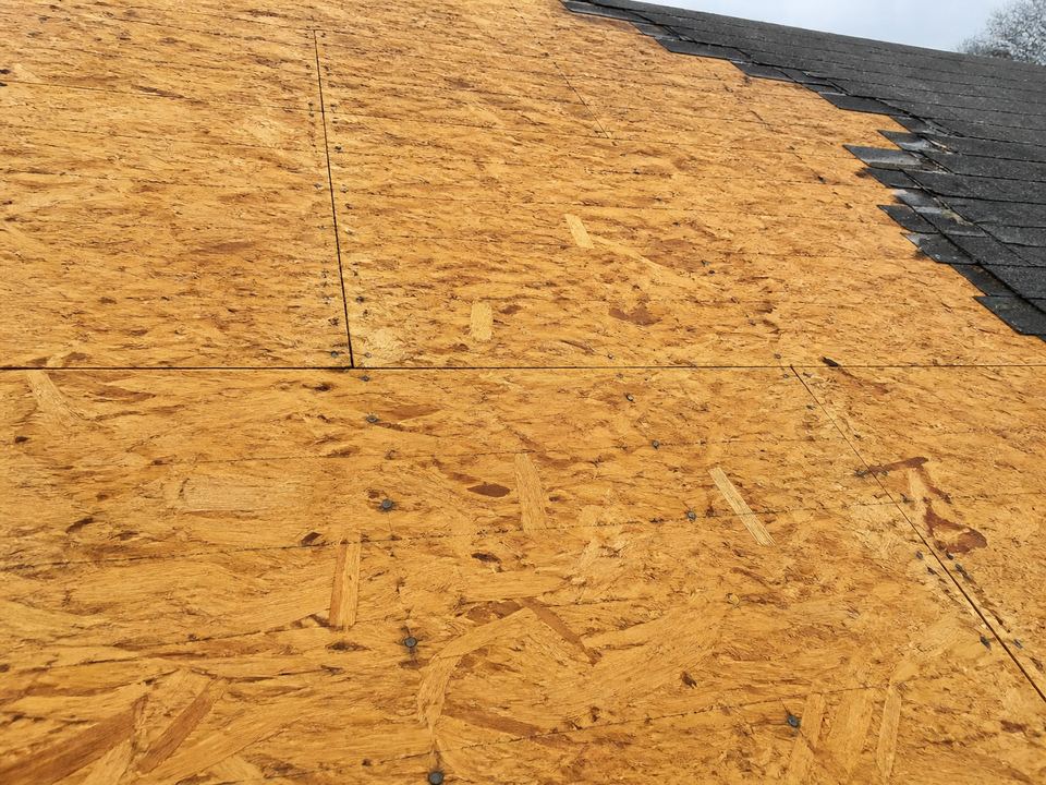 Exposed OSB roof panels with compressed wood strands and panel joints, beside a remaining area of asphalt shingles.