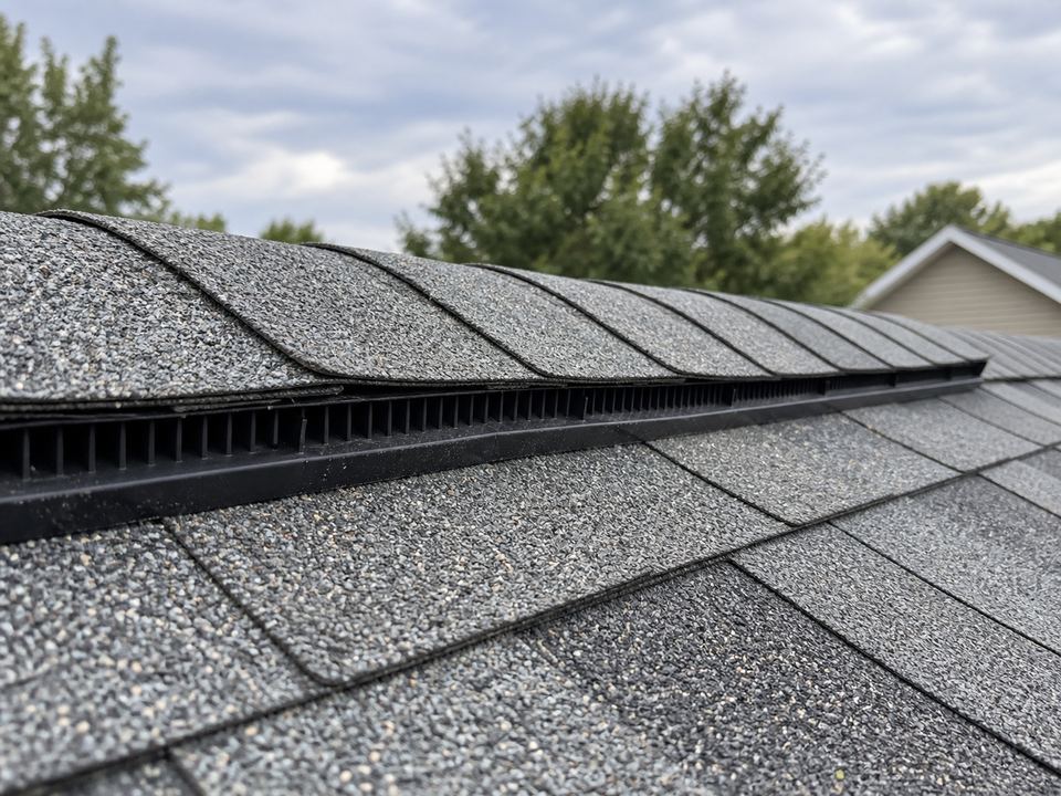 Continuous black vent openings beneath cap shingles along a roof ridge, viewed from the side.