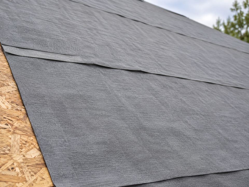 Gray synthetic underlayment sheets overlap across a sloped roof deck, with a narrow strip of OSB visible at the edge.