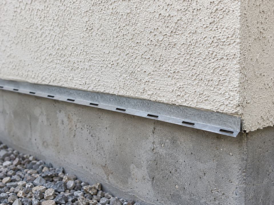 A metal termination strip with open drainage slots runs along the bottom of a stucco wall above exposed concrete foundation.