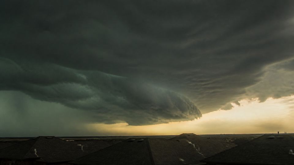 A hard-edged storm shelf cloud advancing over flat North Texas terrain, rooflines a thin band along the bottom.