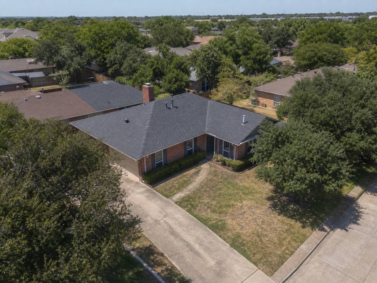 Oblique aerial view of a single-storey red-brick ranch house with an L-shaped plan, roofed in weathered-charcoal architectural shingles, with neighbouring roofs and mature trees around it.