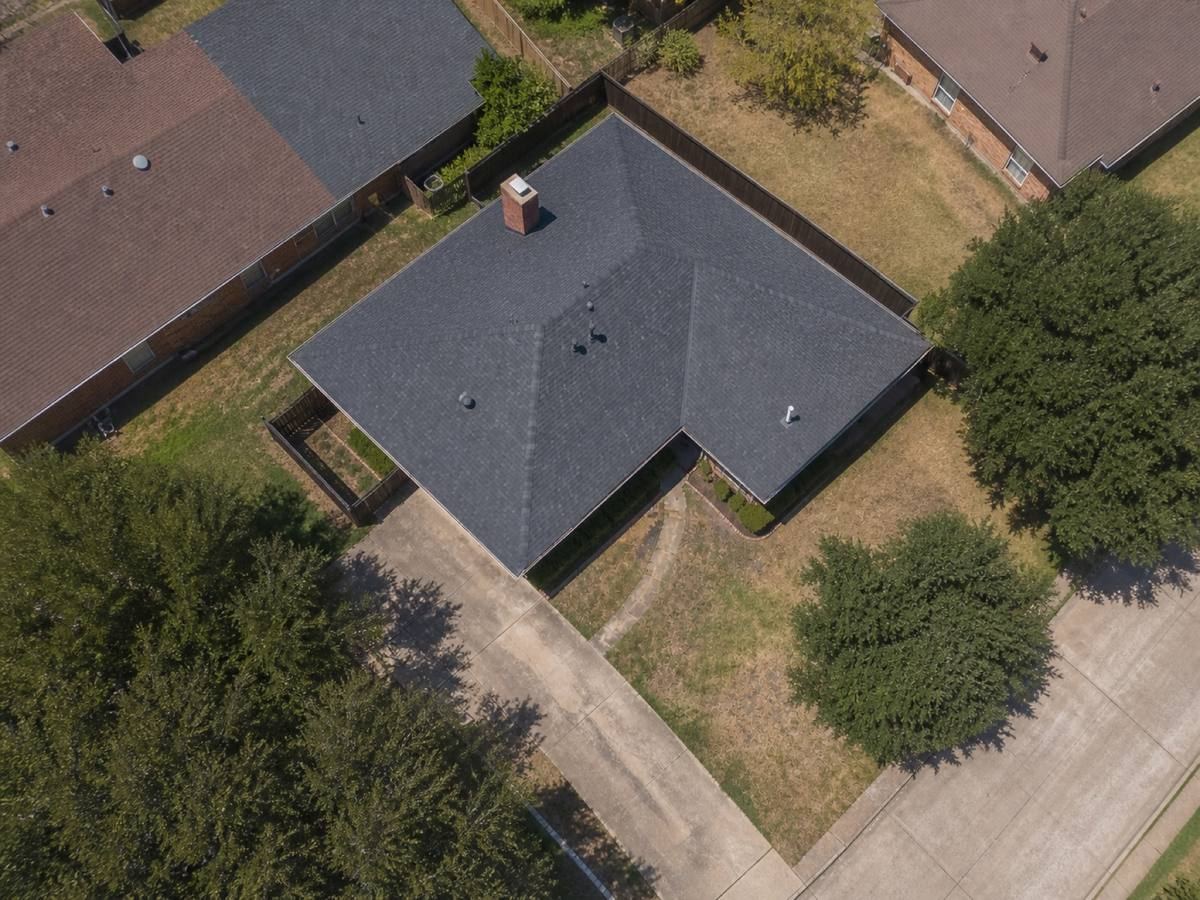 Overhead view looking straight down on a single-storey red-brick ranch house with an L-shaped plan, showing the full run of weathered-charcoal architectural shingles and how the planes drain.