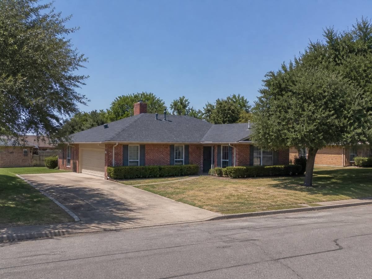 A single-storey red-brick ranch house with an L-shaped plan seen from the street, the weathered-charcoal architectural shingles visible above the elevation.