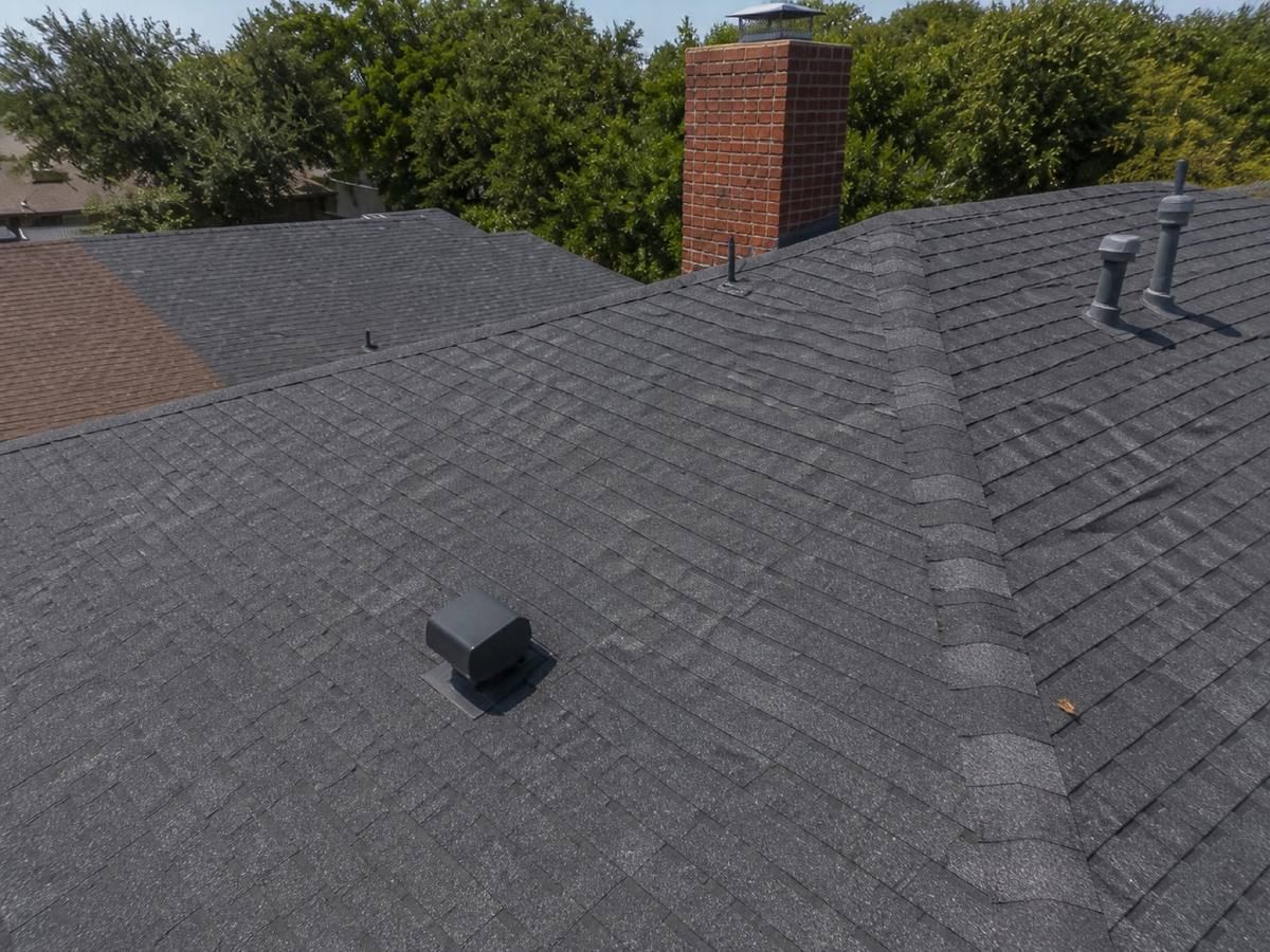 A dark shingle roof meeting at the ridge and hip, with a low box vent, two pipe vents and a red brick chimney behind.