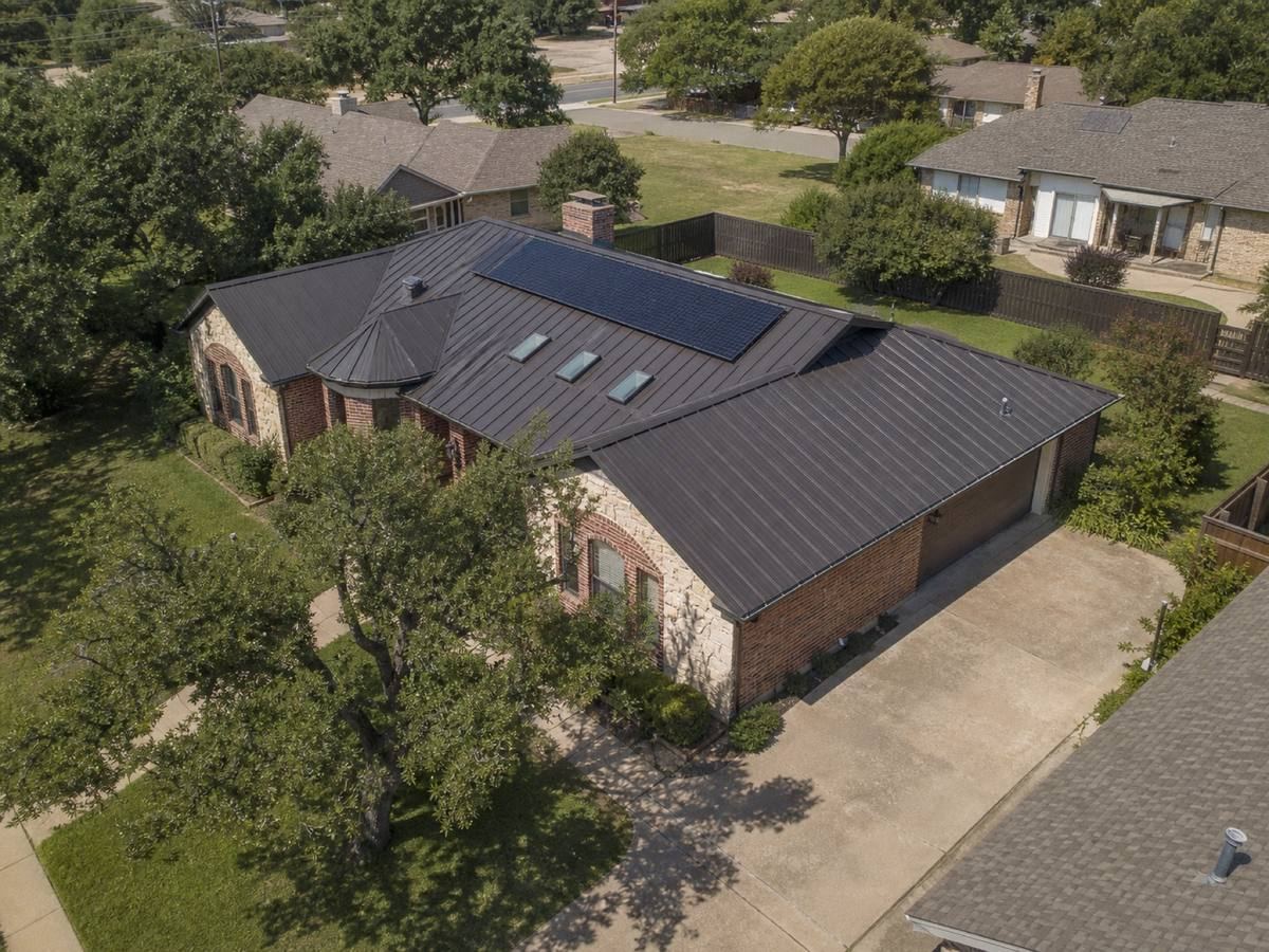Oblique aerial view of a limestone-and-brick single-storey home with shallow cross-gables, roofed in matte bronze standing-seam metal, with neighbouring roofs and mature trees around it.