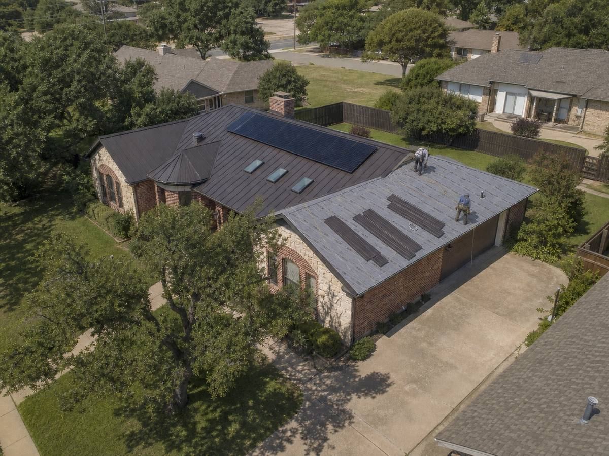A limestone-and-brick single-storey home with shallow cross-gables part-way through a standing-seam metal installation, with finished and unfinished areas both visible.