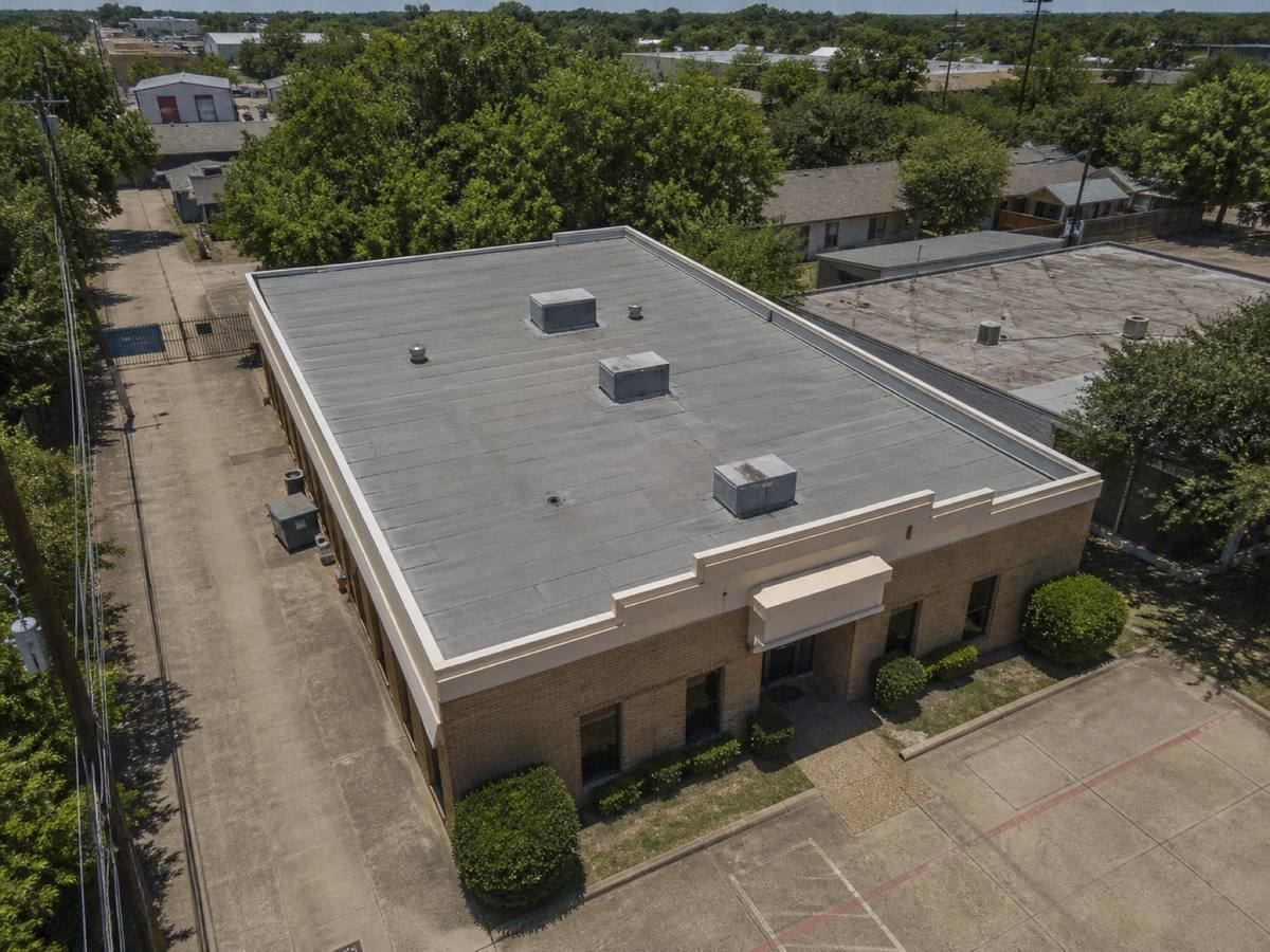 Oblique aerial view of a small two-storey urban commercial building with a cream parapet, roofed in a grey torch-down modified membrane, with neighbouring roofs and mature trees around it.