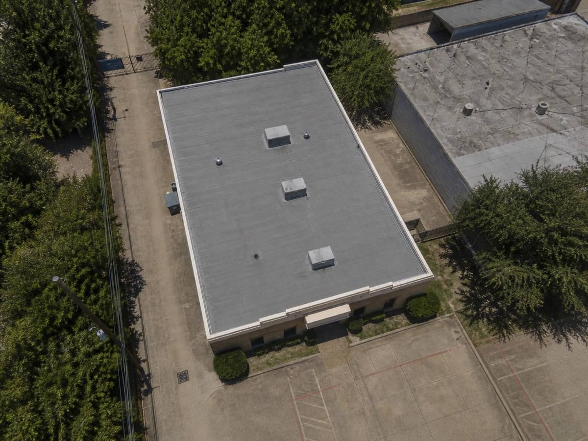 Overhead view looking straight down on a small two-storey urban commercial building with a cream parapet, showing the full run of a grey torch-down modified membrane and how the planes drain.