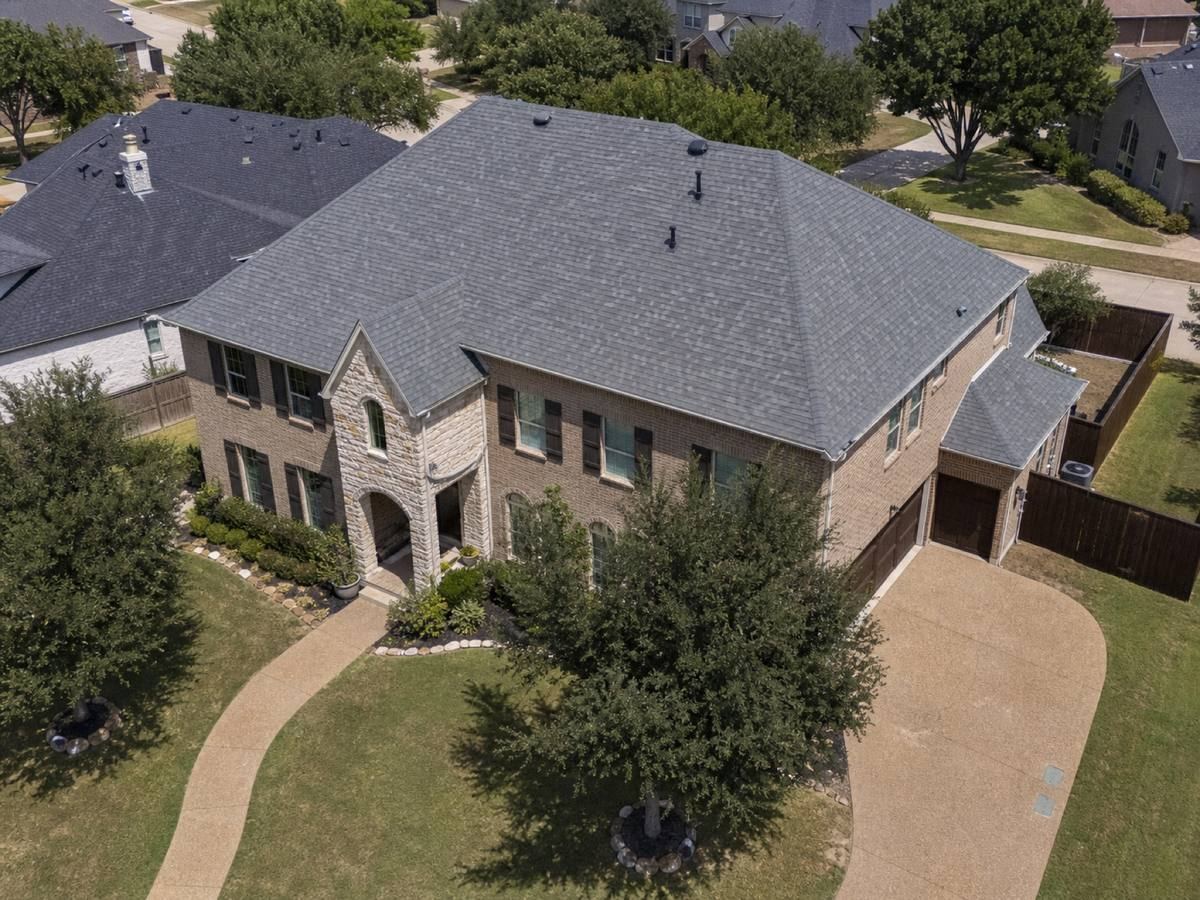 Oblique aerial view of a two-storey tan-brick house with a clipped hip roof and a curved driveway, roofed in slate-grey architectural shingles, with neighbouring roofs and mature trees around it.