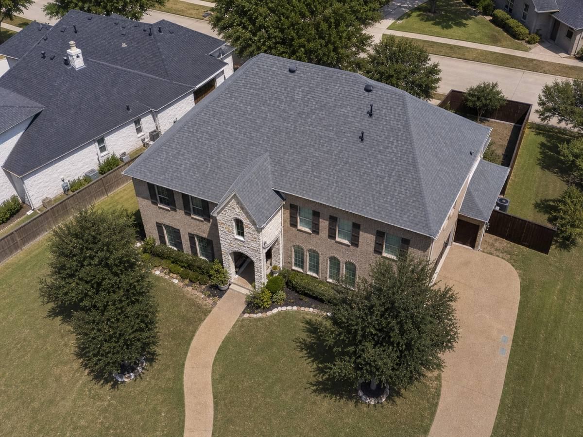 An overhead view of a two-storey brick house showing the whole roof at once — hipped planes, valleys and vent stacks — with the drive and lawn around it.