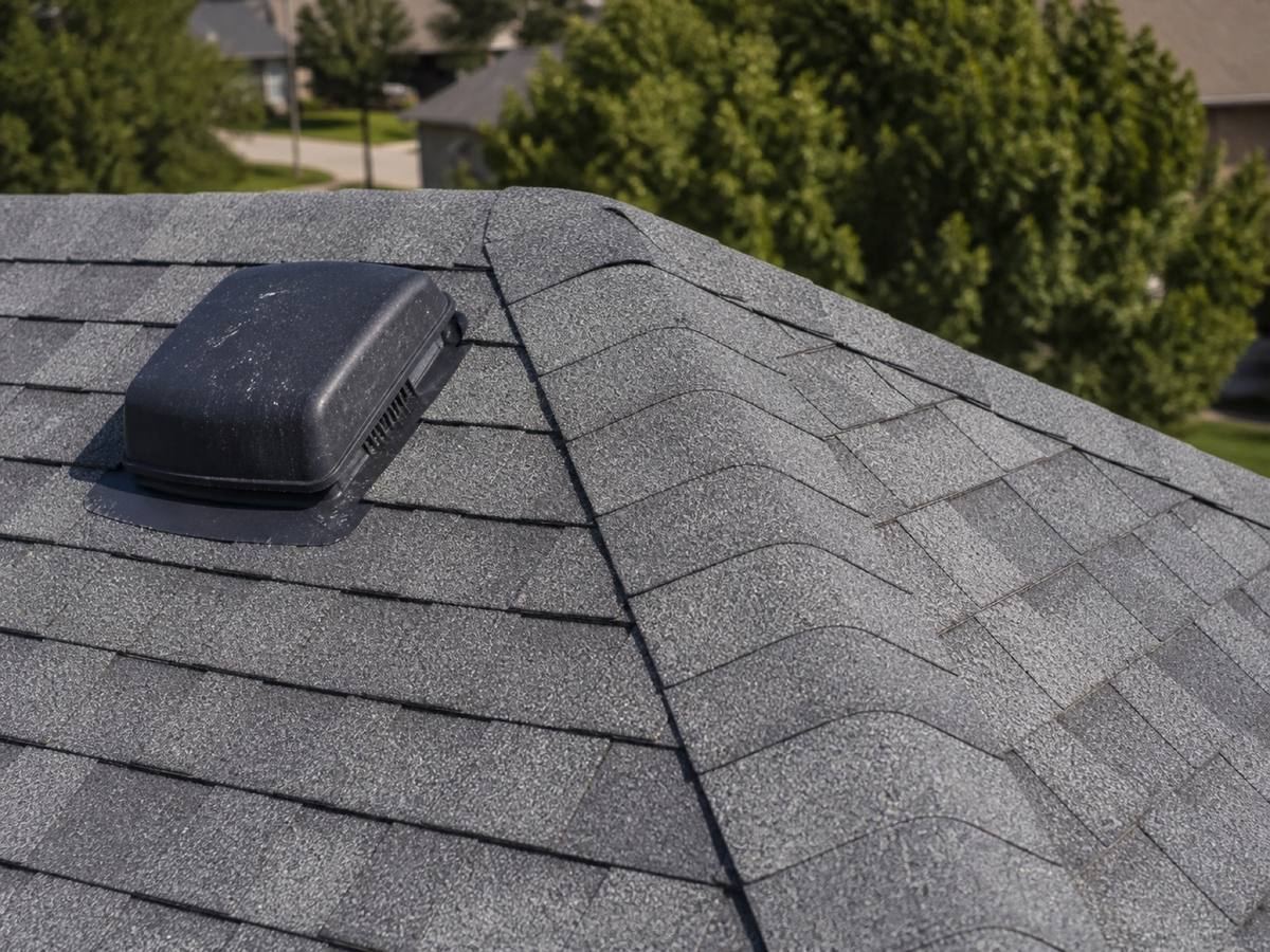 A low black box vent set into a shingle plane near the hip, the courses running away down both slopes behind it.
