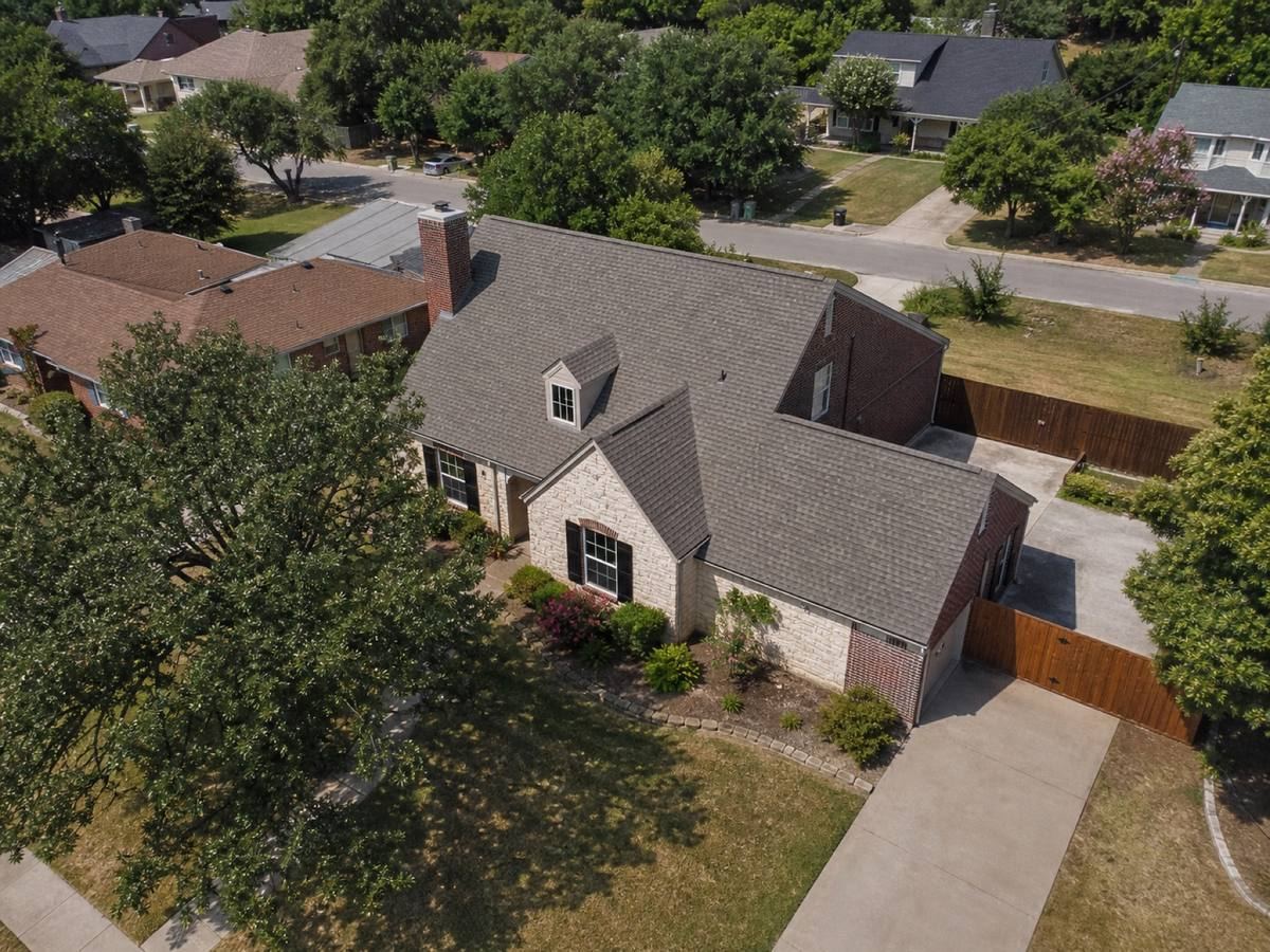 Oblique aerial view of a one-and-a-half-storey house with a stone frontage and an offset garage, roofed in warm-grey impact-resistant shingles, with neighbouring roofs and mature trees around it.
