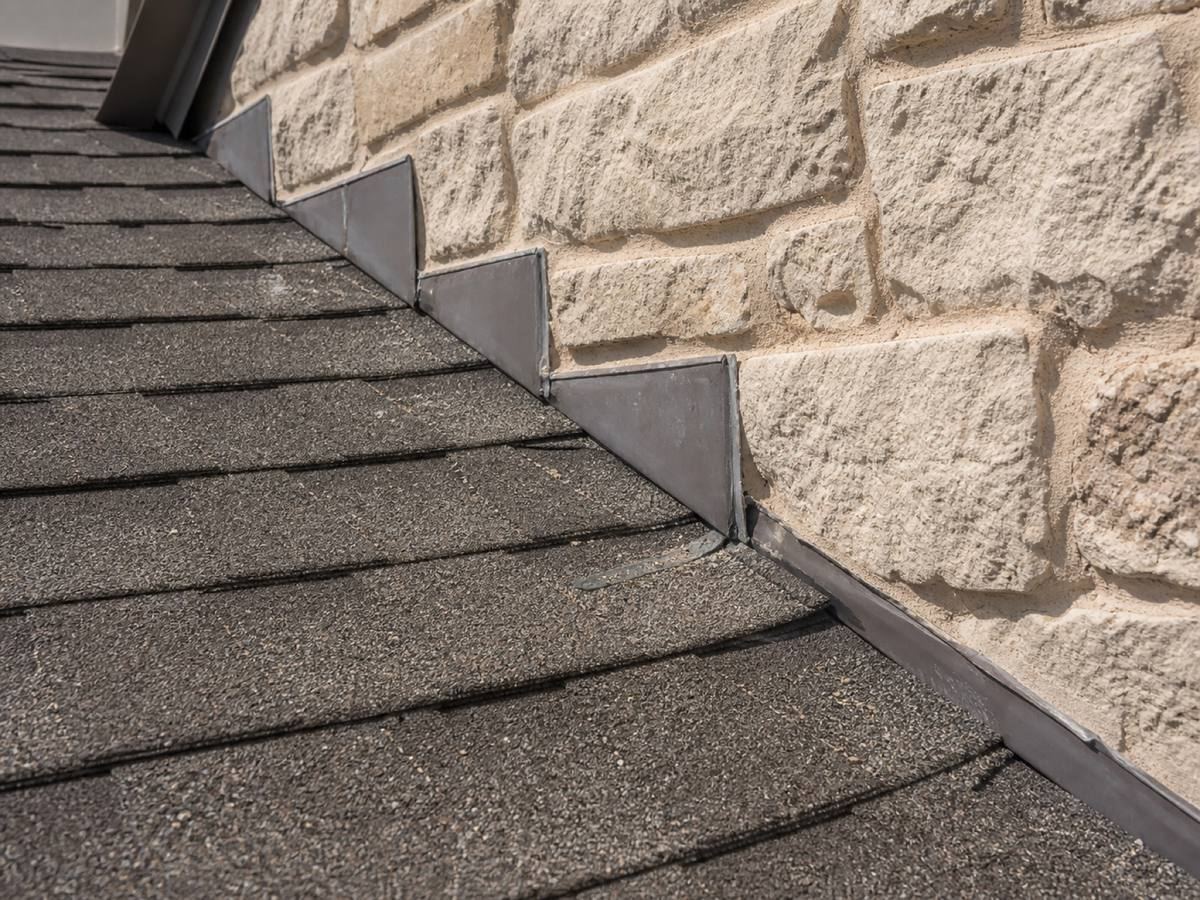 Individual metal step flashings stepping up a rough limestone wall where a shingle plane meets it, the shingle courses running away below.