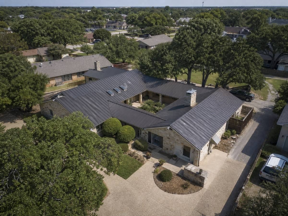 Oblique aerial view of a long single-storey stone house arranged around a courtyard, roofed in matte charcoal standing-seam metal, with neighbouring roofs and mature trees around it.