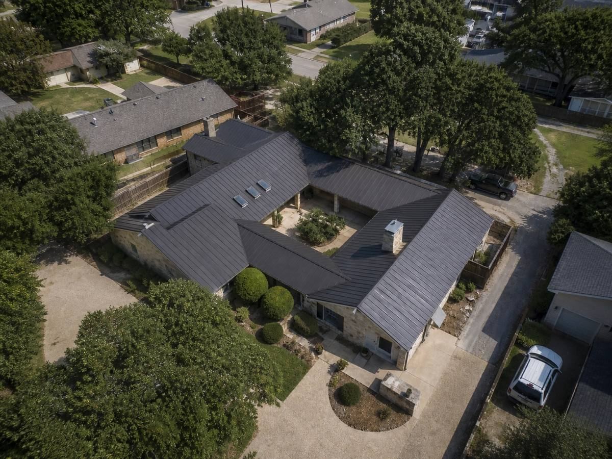 Overhead view looking straight down on a long single-storey stone house arranged around a courtyard, showing the full run of matte charcoal standing-seam metal and how the planes drain.