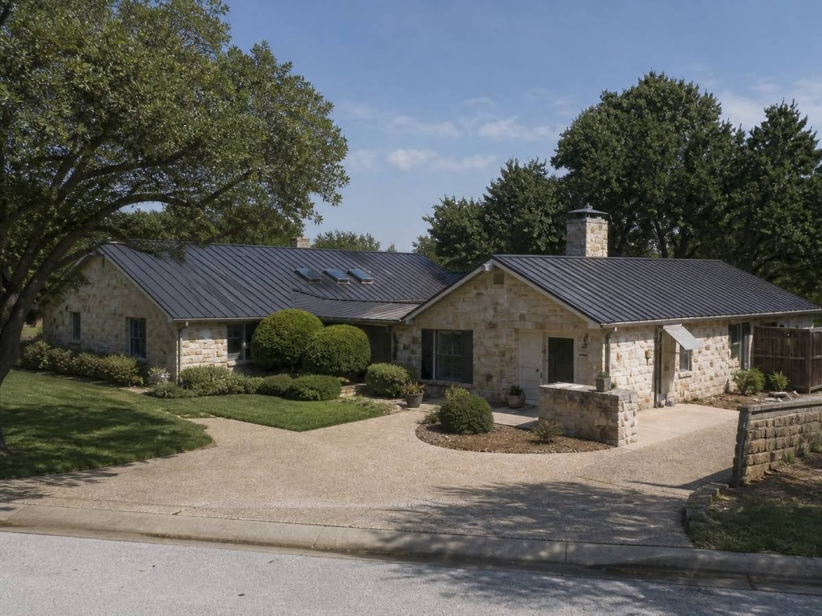 A long single-storey stone house arranged around a courtyard seen from the street, the matte charcoal standing-seam metal visible above the elevation.
