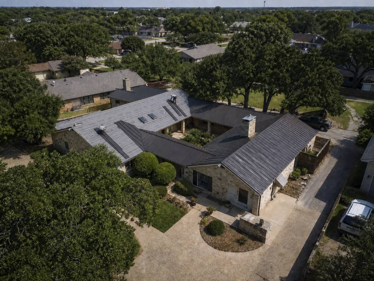 A long single-storey stone house arranged around a courtyard part-way through a standing-seam metal installation, with finished and unfinished areas both visible.