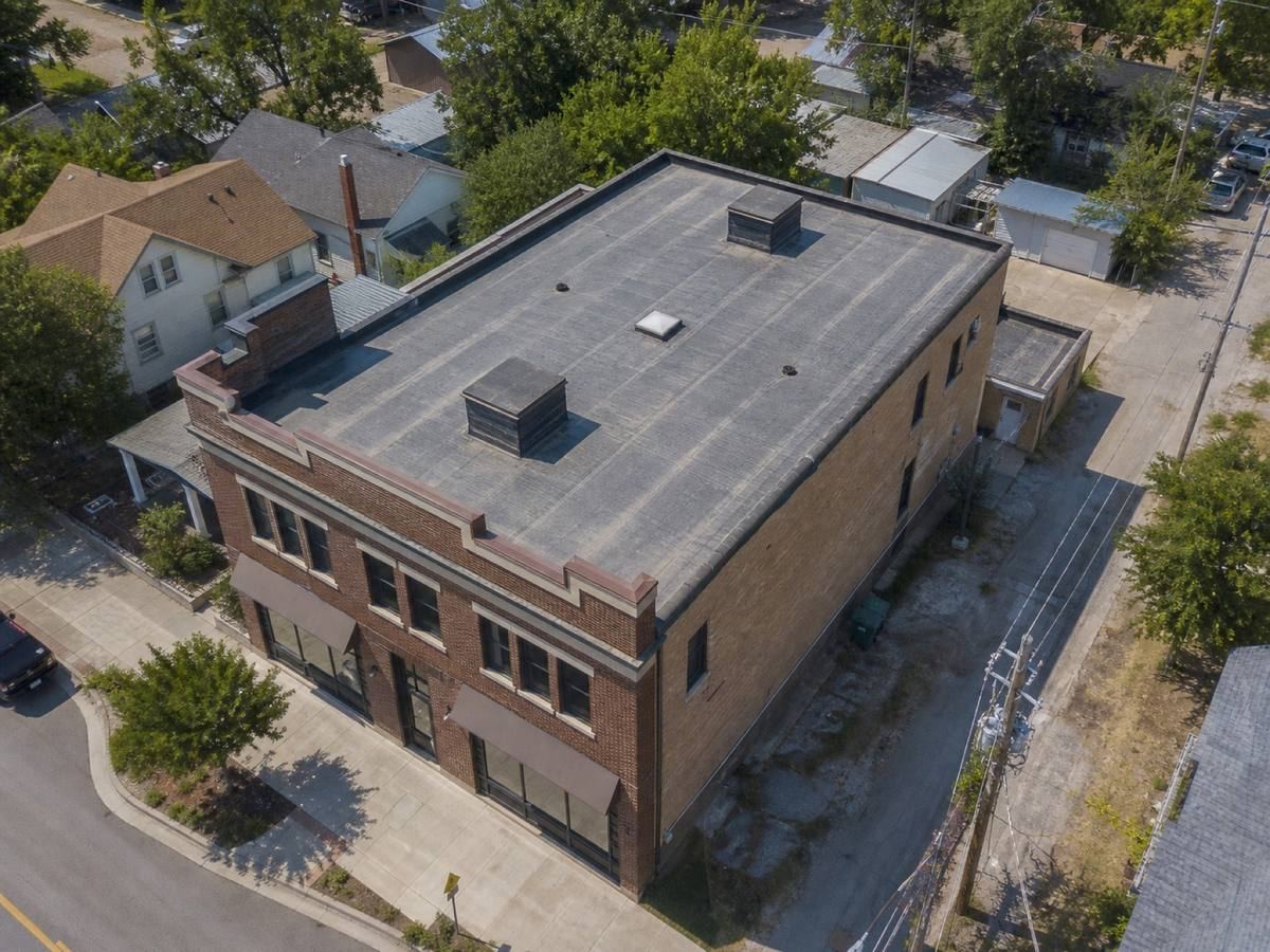Oblique aerial view of a mixed-use brick building with a stepped parapet, roofed in a dark-grey low-slope membrane, with neighbouring roofs and mature trees around it.