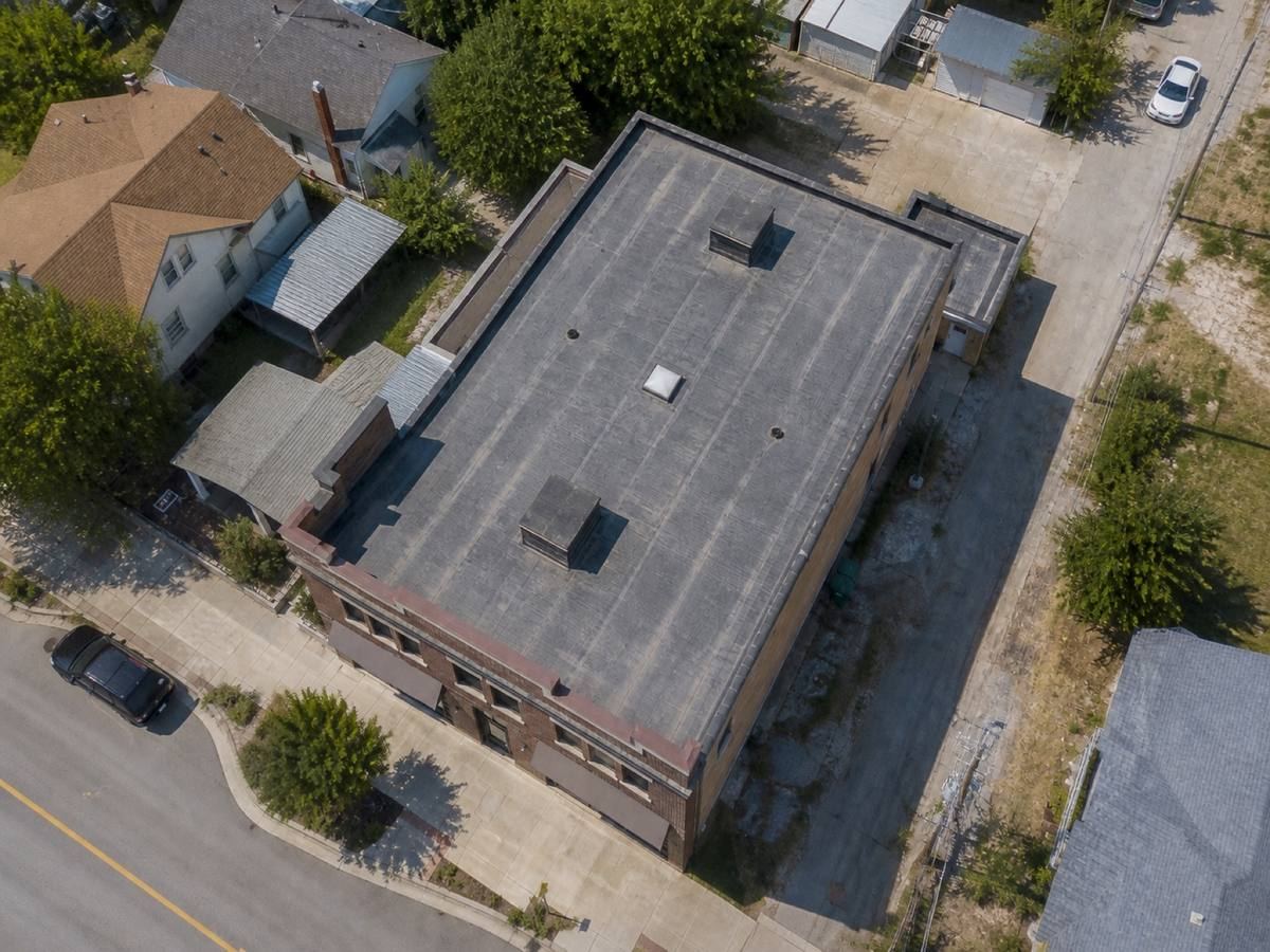 Overhead view looking straight down on a mixed-use brick building with a stepped parapet, showing the full run of a dark-grey low-slope membrane and how the planes drain.