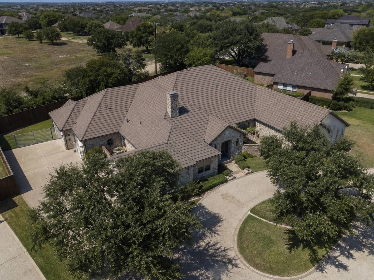 Oblique aerial view of a broad single-storey stone residence with irregular hips and a circular drive, roofed in muted brown concrete tile, with neighbouring roofs and mature trees around it.