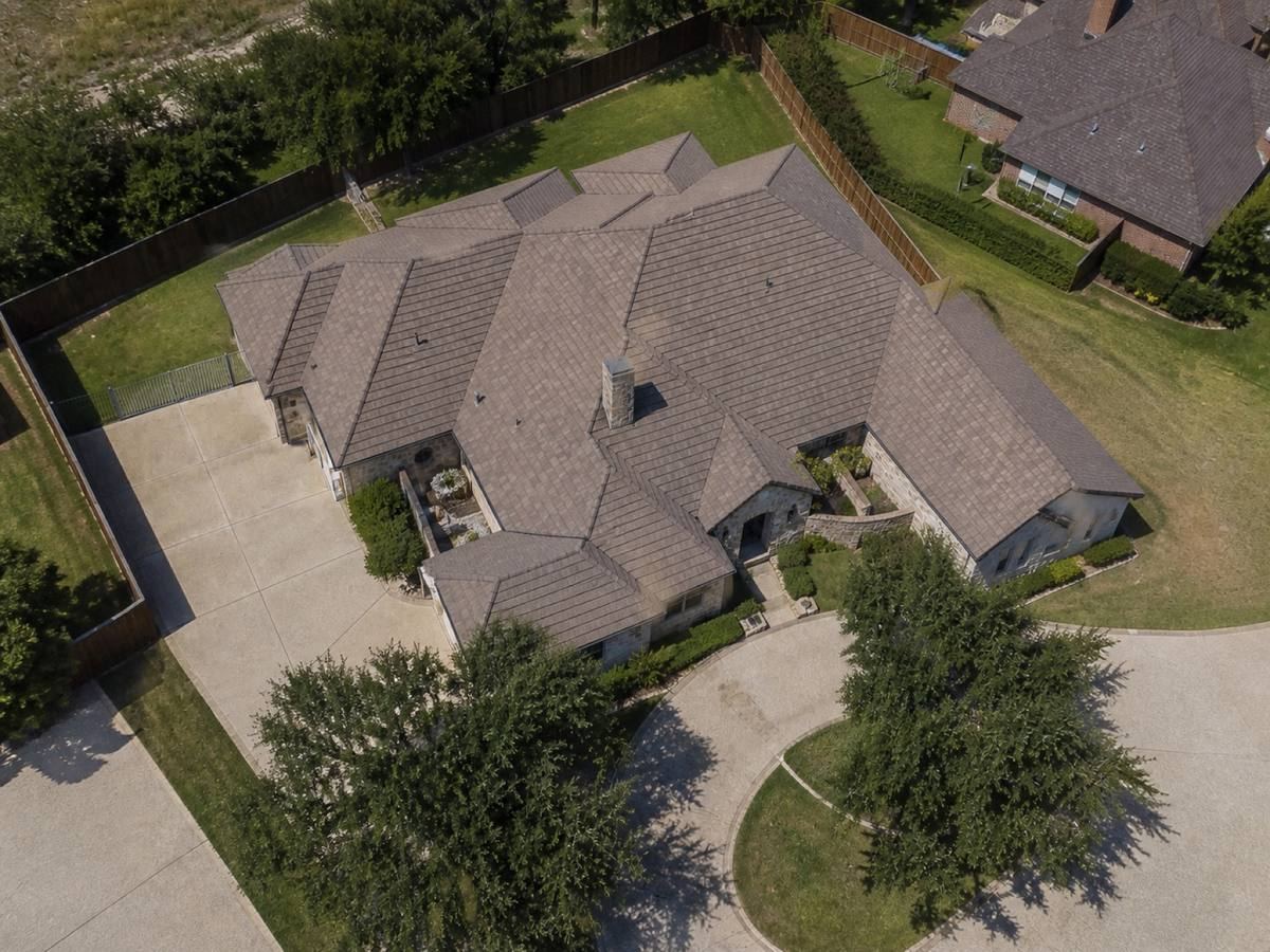 Overhead view looking straight down on a broad single-storey stone residence with irregular hips and a circular drive, showing the full run of muted brown concrete tile and how the planes drain.