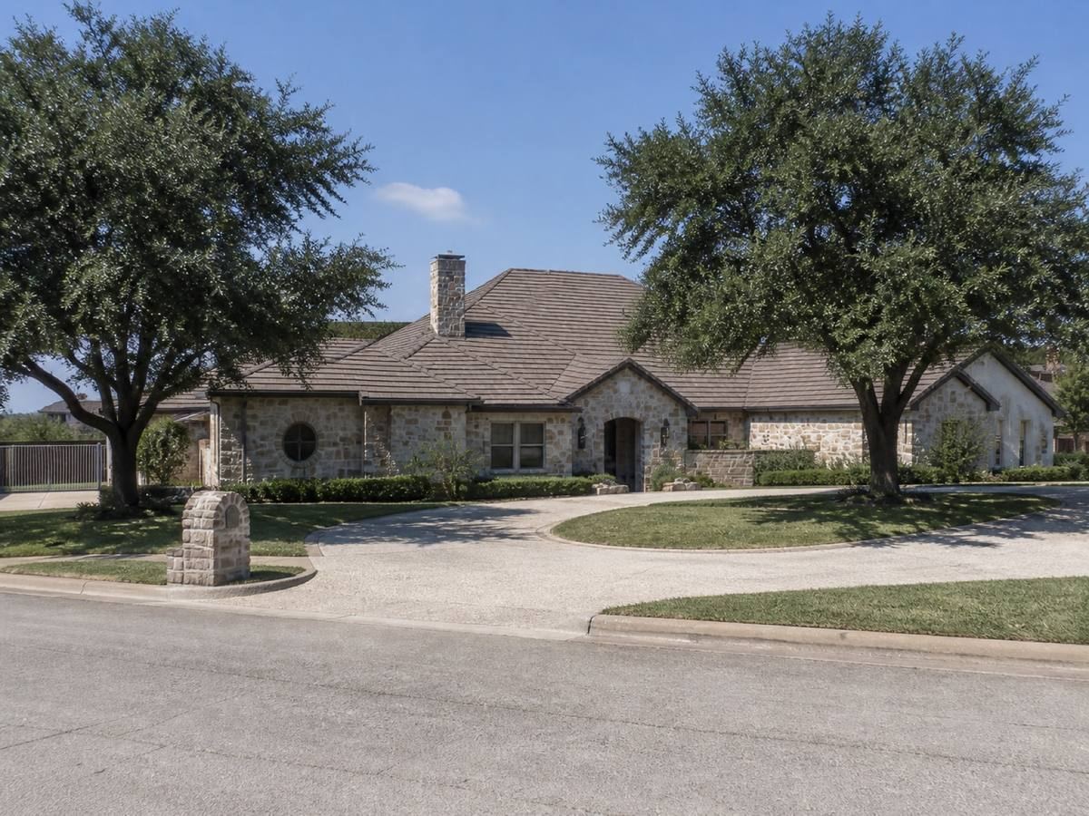 A broad single-storey stone residence with irregular hips and a circular drive seen from the street, the muted brown concrete tile visible above the elevation.