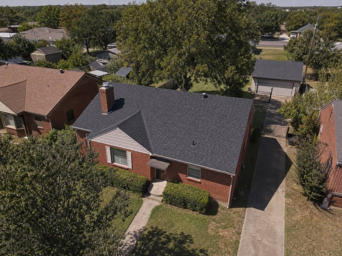 Oblique aerial view of a modest red-brick bungalow on a narrow lot with a detached rear garage, roofed in charcoal dimensional shingles, with neighbouring roofs and mature trees around it.