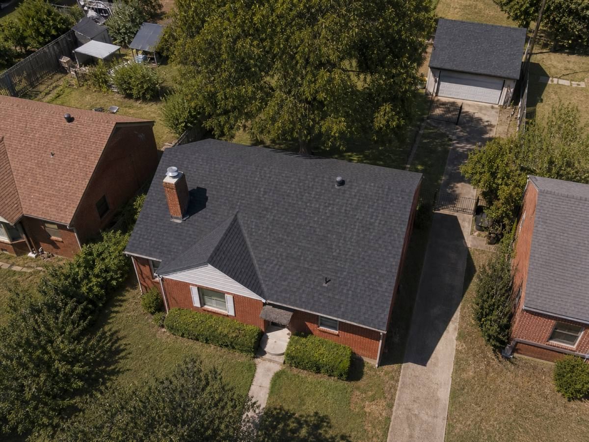 Overhead view looking straight down on a modest red-brick bungalow on a narrow lot with a detached rear garage, showing the full run of charcoal dimensional shingles and how the planes drain.