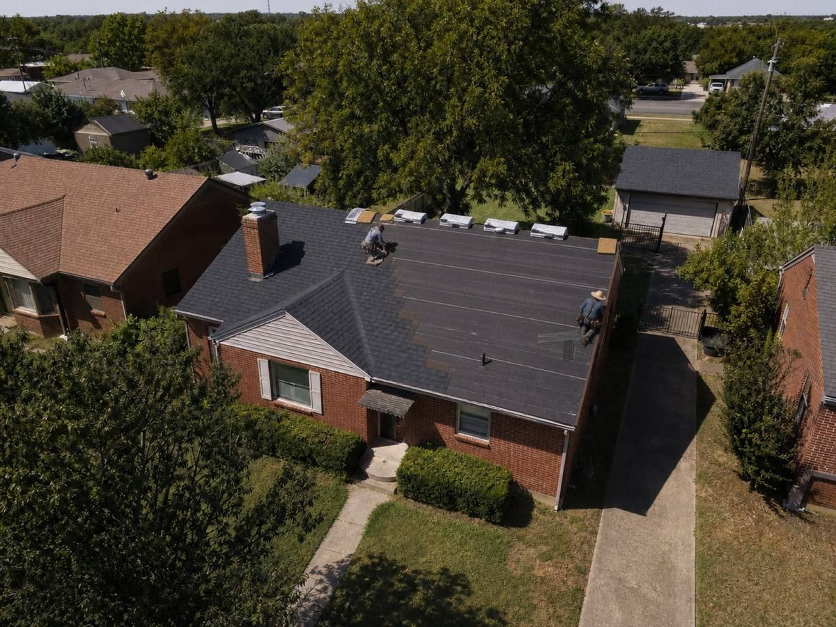 A modest red-brick bungalow on a narrow lot with a detached rear garage part-way through a architectural shingle installation, with finished and unfinished areas both visible.