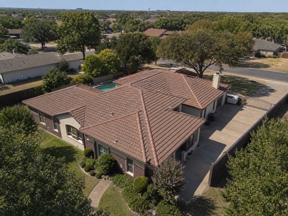 Oblique aerial view of a cream stucco-and-brick house with low hips and a rear pool, roofed in weathered terracotta concrete tile, with neighbouring roofs and mature trees around it.