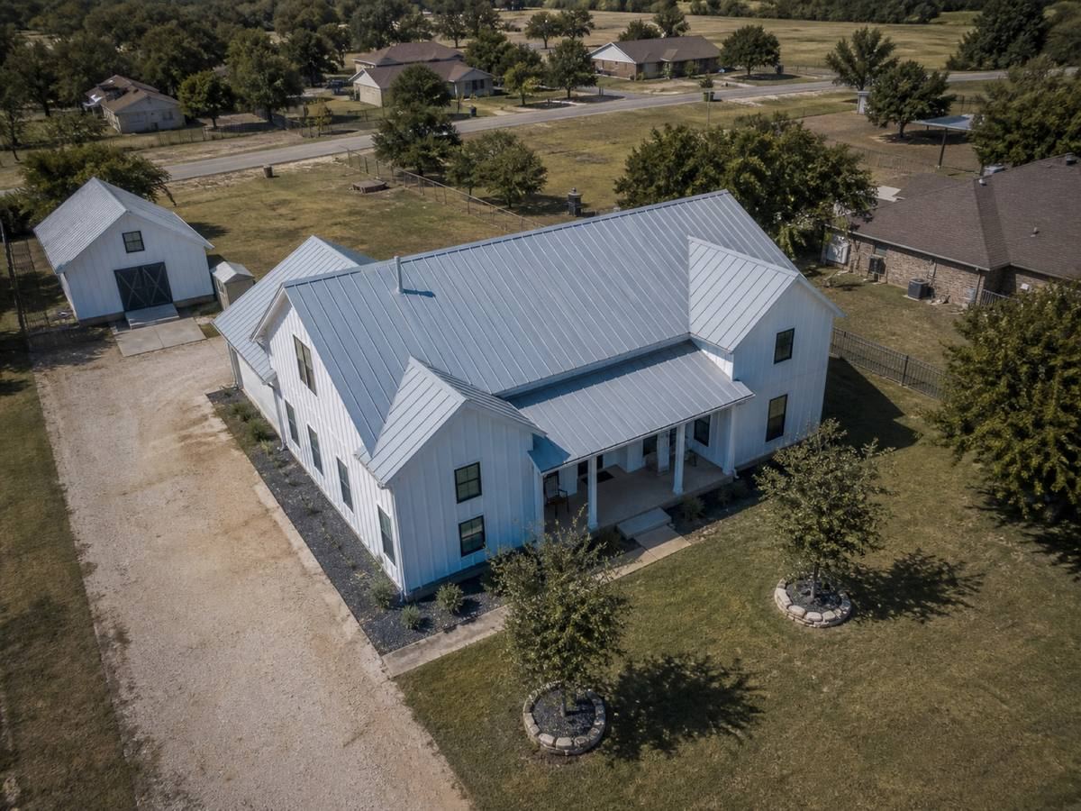 Oblique aerial view of a contemporary white board-and-batten farmhouse with black window trim, roofed in Galvalume standing-seam metal, with neighbouring roofs and mature trees around it.