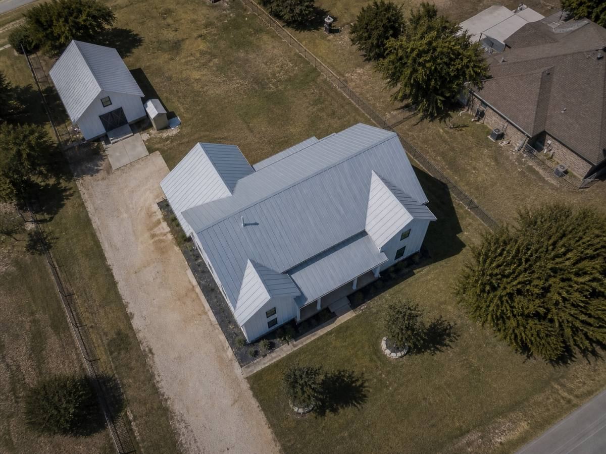 Overhead view looking straight down on a contemporary white board-and-batten farmhouse with black window trim, showing the full run of Galvalume standing-seam metal and how the planes drain.