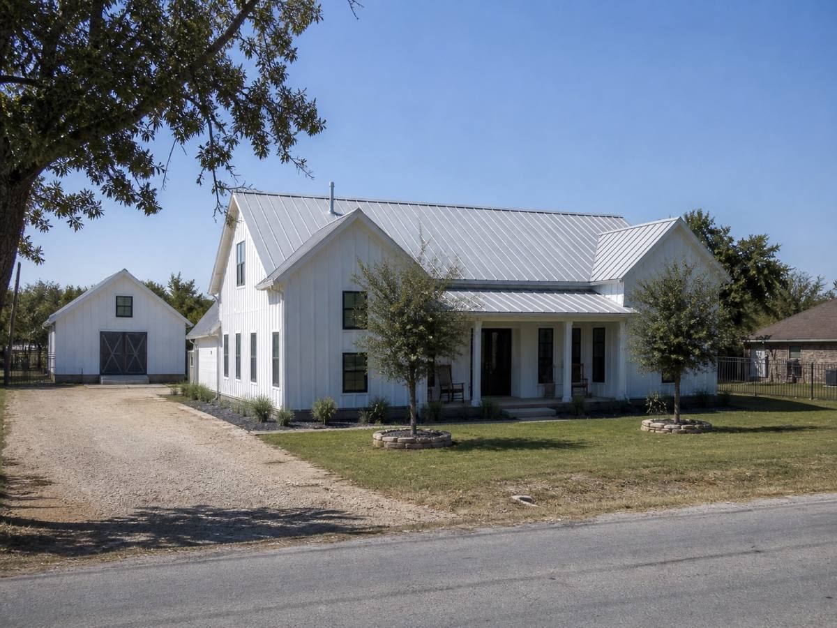 A contemporary white board-and-batten farmhouse with black window trim seen from the street, the Galvalume standing-seam metal visible above the elevation.