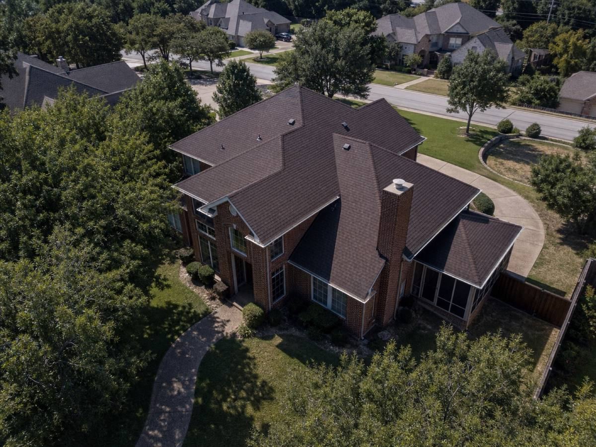 Oblique aerial view of a large brick house with multiple hips and a screened rear porch, roofed in deep-brown architectural shingles, with neighbouring roofs and mature trees around it.