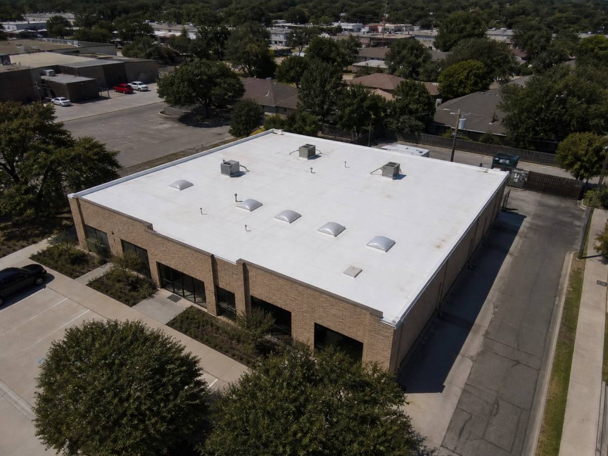 Oblique aerial view of a single-storey brick retail building with a rectangular roof, roofed in a white TPO membrane, with neighbouring roofs and mature trees around it.