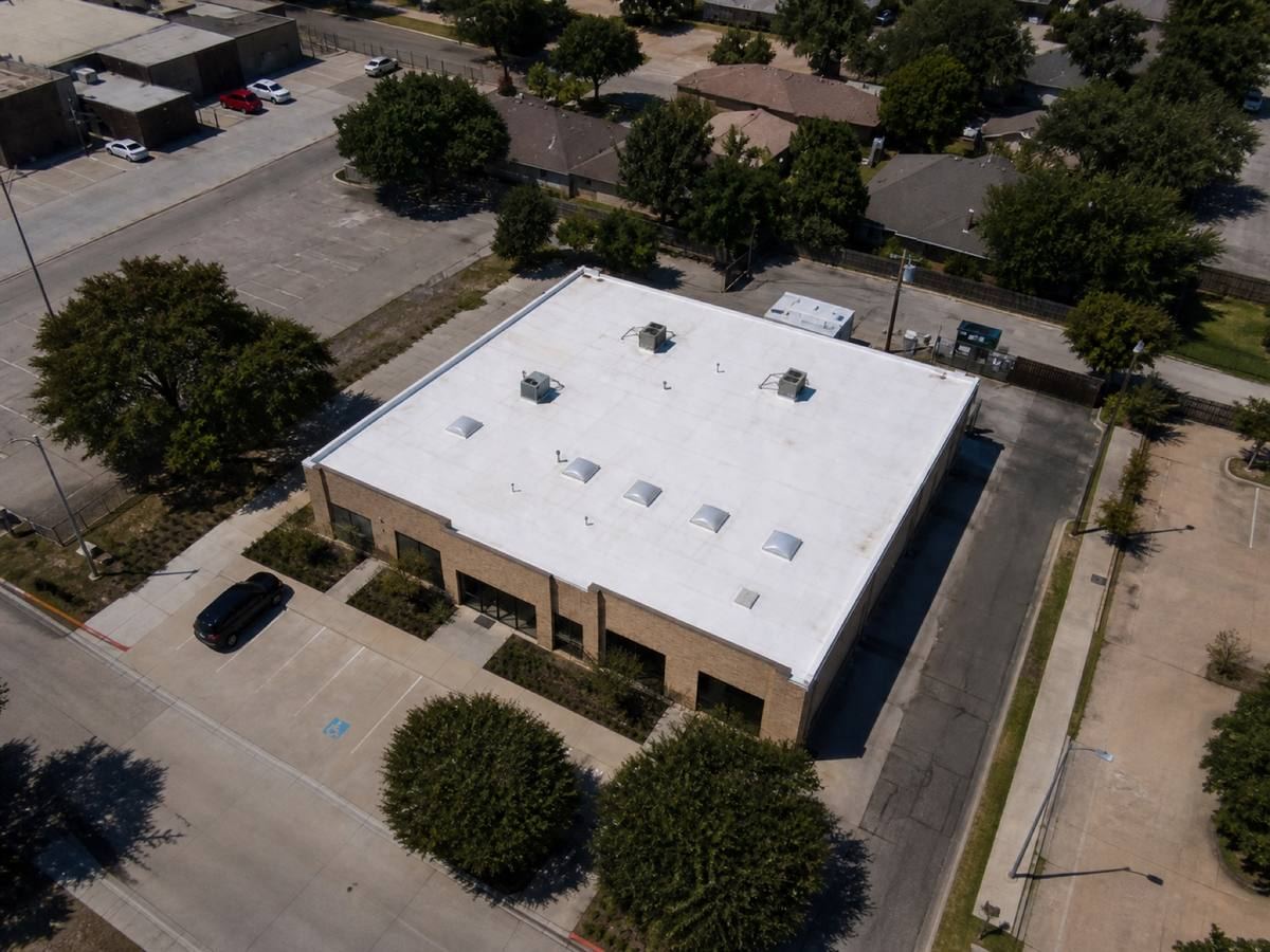 Overhead view looking straight down on a single-storey brick retail building with a rectangular roof, showing the full run of a white TPO membrane and how the planes drain.