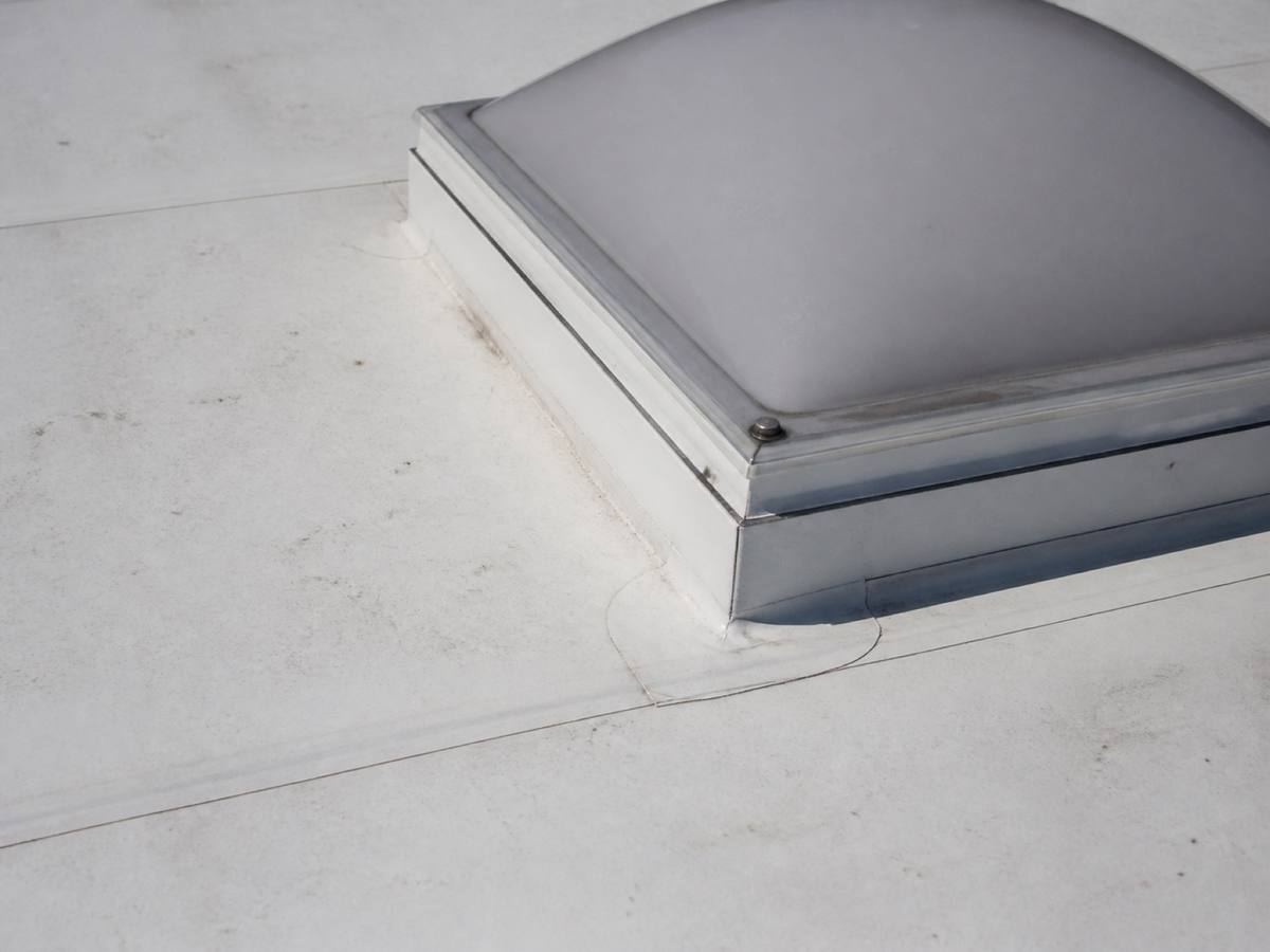 A domed rooflight on a raised kerb in a white single-ply membrane roof, welded seams running across the field around it.