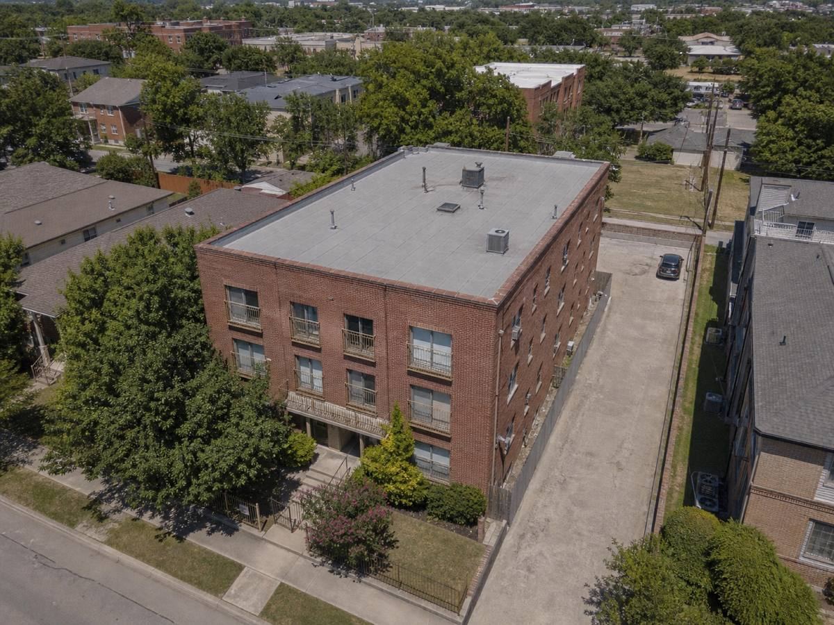 Oblique aerial view of a compact brick apartment building with parapets on all sides, roofed in a medium-grey modified-bitumen cap sheet, with neighbouring roofs and mature trees around it.