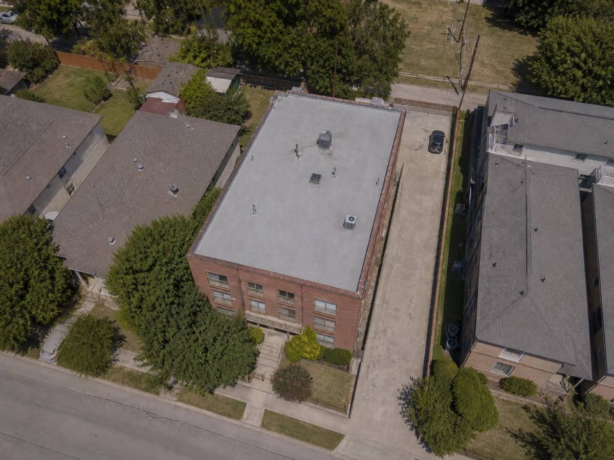 Overhead view looking straight down on a compact brick apartment building with parapets on all sides, showing the full run of a medium-grey modified-bitumen cap sheet and how the planes drain.