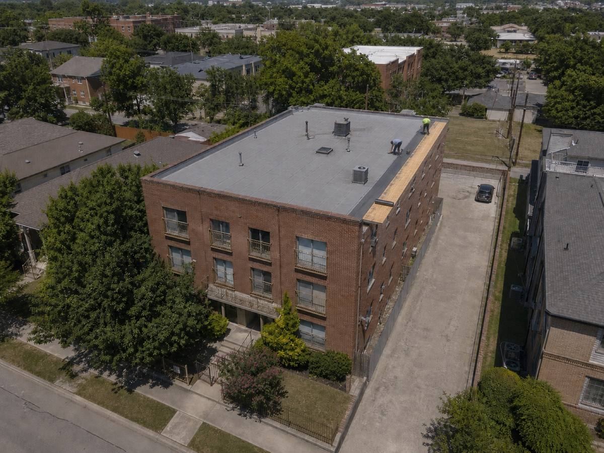 A compact brick apartment building with parapets on all sides part-way through a modified bitumen installation, with finished and unfinished areas both visible.