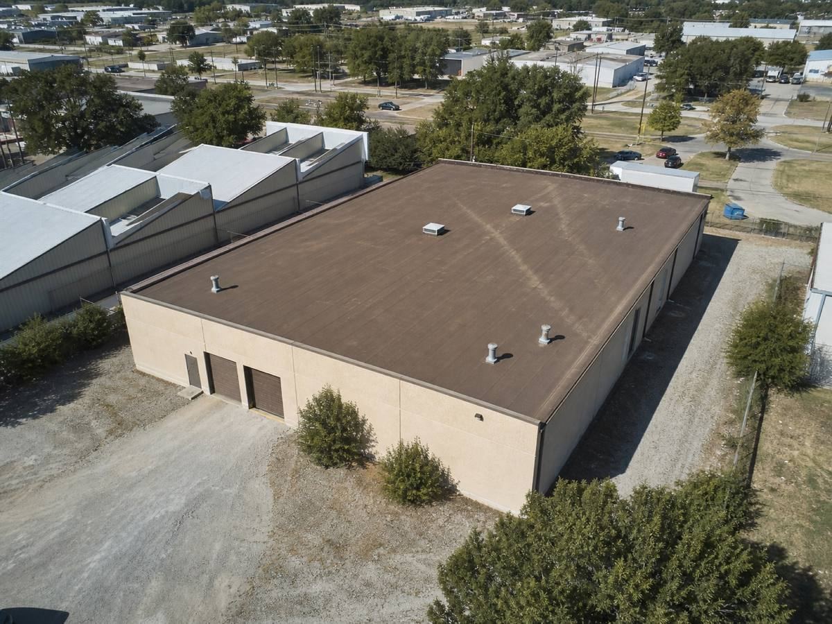Oblique aerial view of a small warehouse with a gravel service yard and sawtooth roofs behind it, roofed in a brown mineral-surface cap sheet, with neighbouring roofs and mature trees around it.