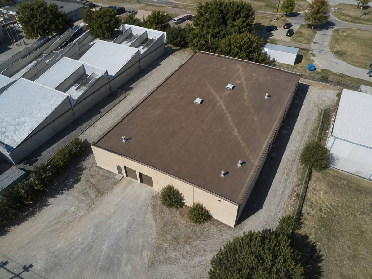 Overhead view looking straight down on a small warehouse with a gravel service yard and sawtooth roofs behind it, showing the full run of a brown mineral-surface cap sheet and how the planes drain.
