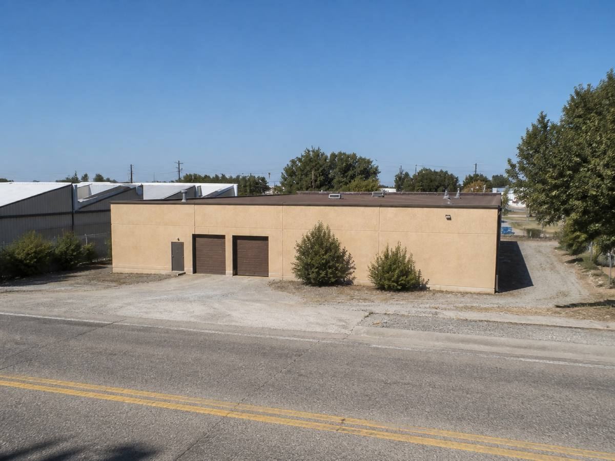 A small warehouse with a gravel service yard and sawtooth roofs behind it seen from the street, the a brown mineral-surface cap sheet visible above the elevation.