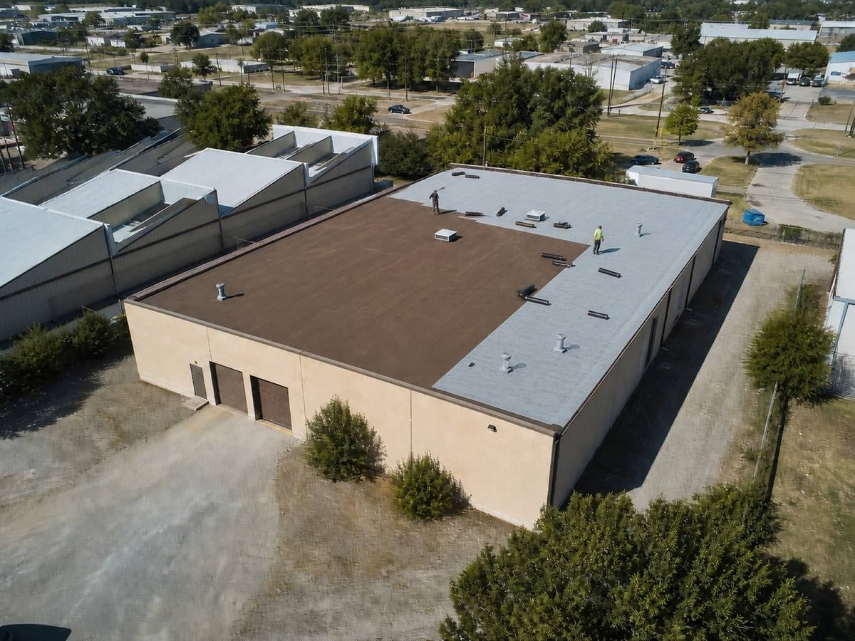 A small warehouse with a gravel service yard and sawtooth roofs behind it part-way through a modified bitumen installation, with finished and unfinished areas both visible.
