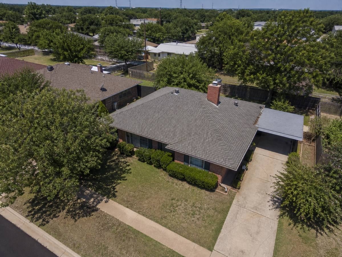 Oblique aerial view of a 1970s single-storey brick house with a simple hip roof and a side carport, roofed in cool-grey architectural shingles, with neighbouring roofs and mature trees around it.