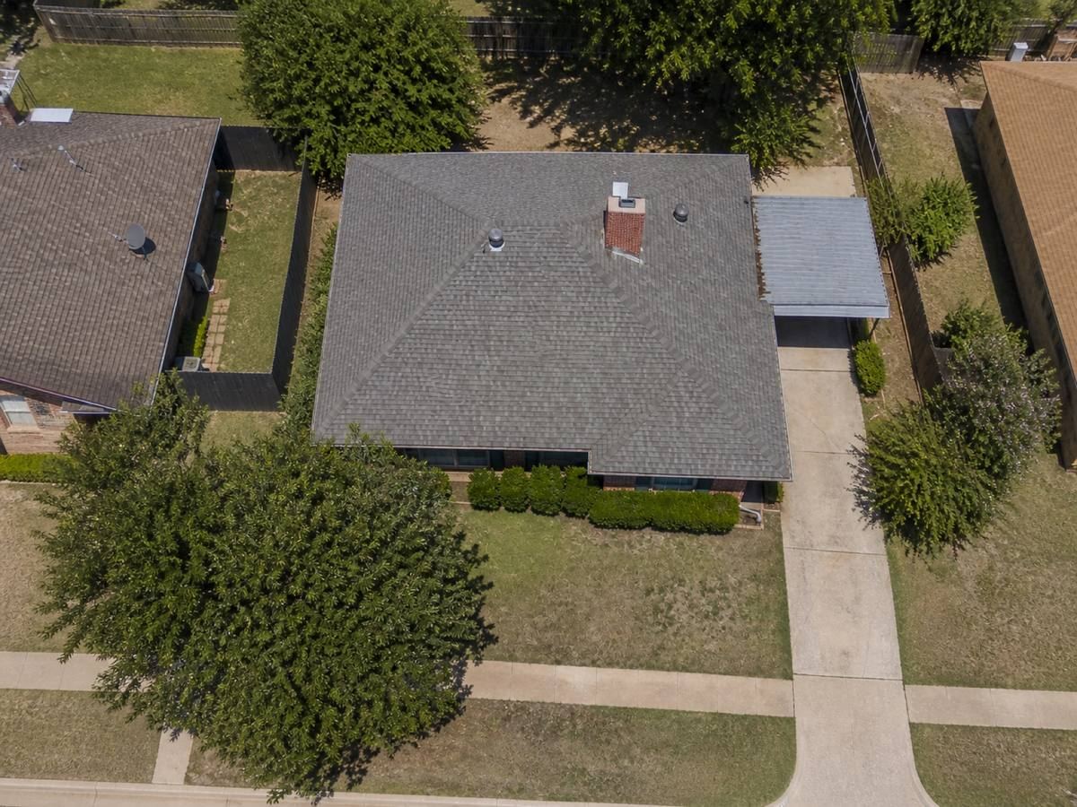 Overhead view looking straight down on a 1970s single-storey brick house with a simple hip roof and a side carport, showing the full run of cool-grey architectural shingles and how the planes drain.