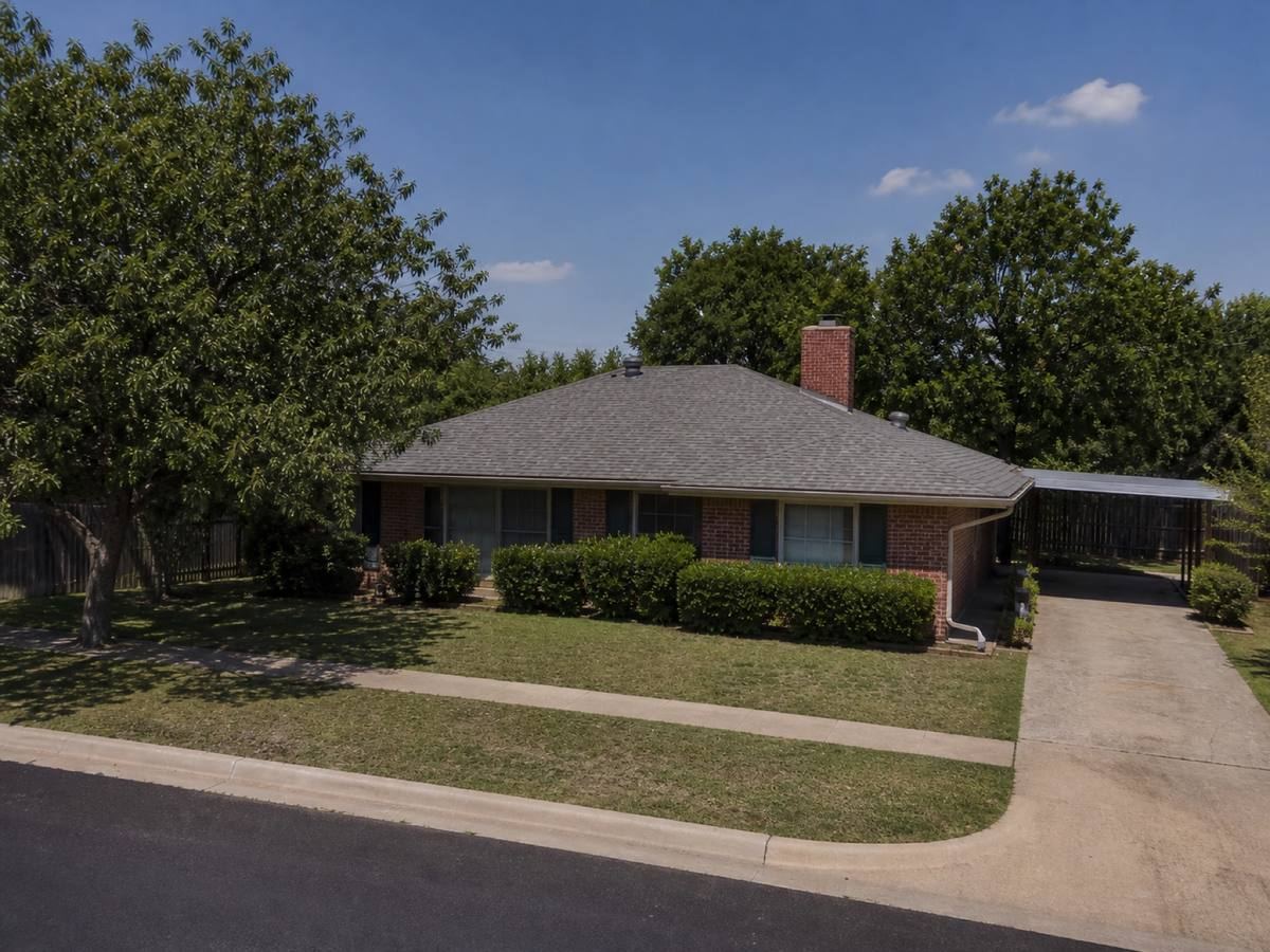 A 1970s single-storey brick house with a simple hip roof and a side carport seen from the street, the cool-grey architectural shingles visible above the elevation.