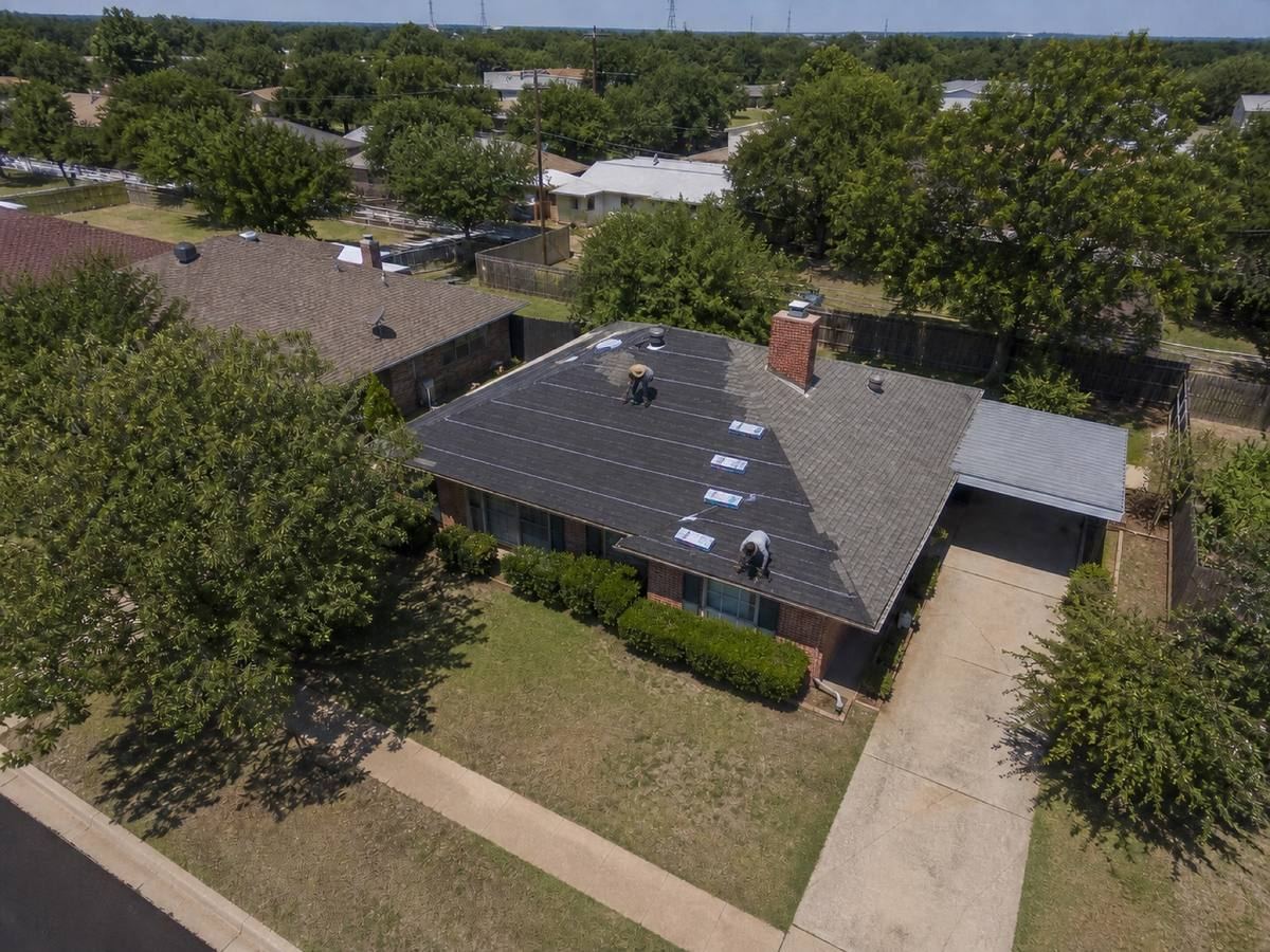 A 1970s single-storey brick house with a simple hip roof and a side carport part-way through a architectural shingle installation, with finished and unfinished areas both visible.